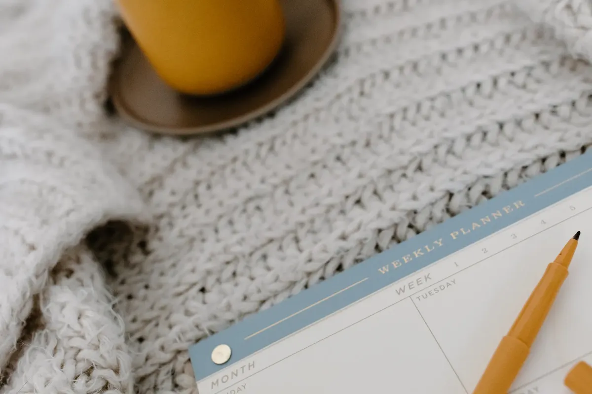 A cozy flat lay setup with a weekly planner, pen, and warm drink on a knitted blanket.